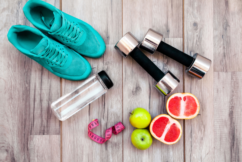Workout flat lay with turquoise sneakers, dumbbells, water, measuring tape, apples, and sliced grapefruits on wood.