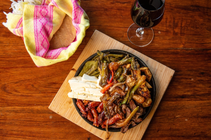 A cast iron skillet of chorizo, sliced cheese, peppers, and onions on a wooden board, with red wine.