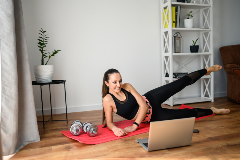 Woman in black workout clothes exercises on a red mat, watching a laptop, with weights nearby.