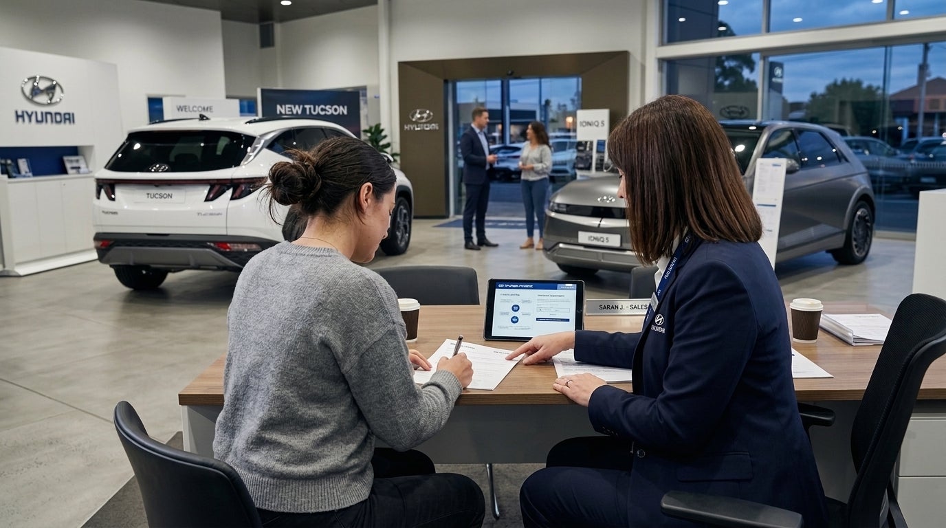 A woman signs paperwork at a desk with a sales representative inside a Hyundai car dealership showroom. (Representative image for illustration purposes only. Actual vehicle may vary based on trim and configuration.)