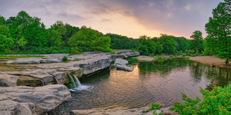 Panoramic view of a calm river with small waterfalls over flat rock formations, surrounded by lush green trees at sunset.