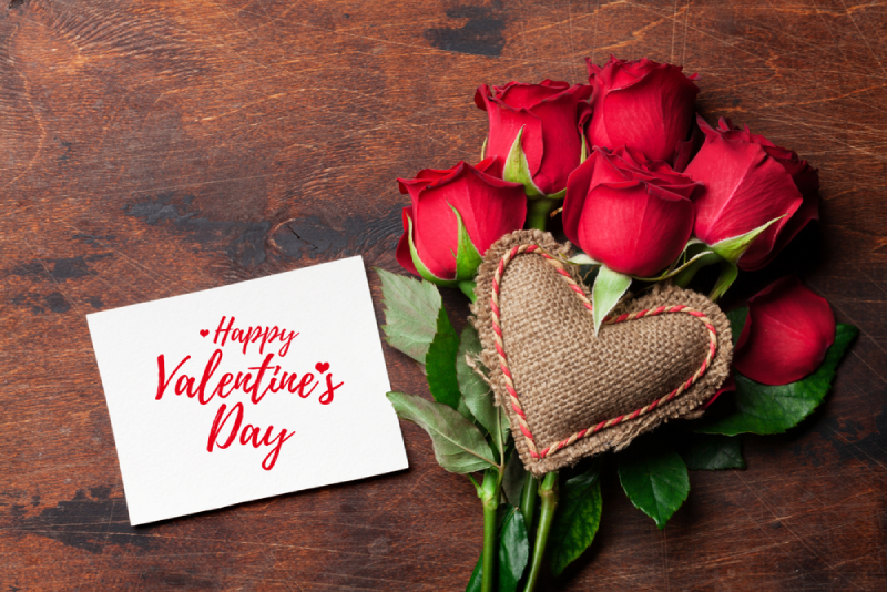 A "Happy Valentine's Day" card next to a bouquet of red roses and a burlap heart on a dark wooden table.