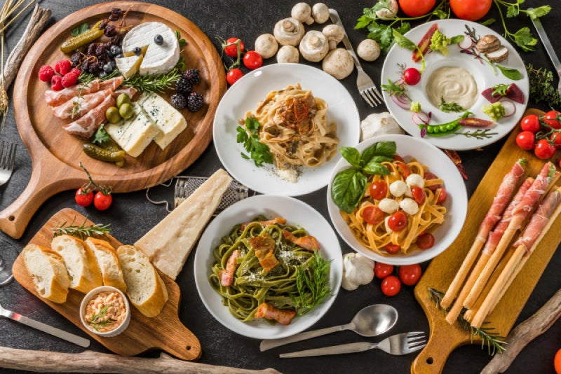 A rich assortment of Italian dishes, charcuterie, cheeses, and fresh ingredients artfully arranged on a dark surface.