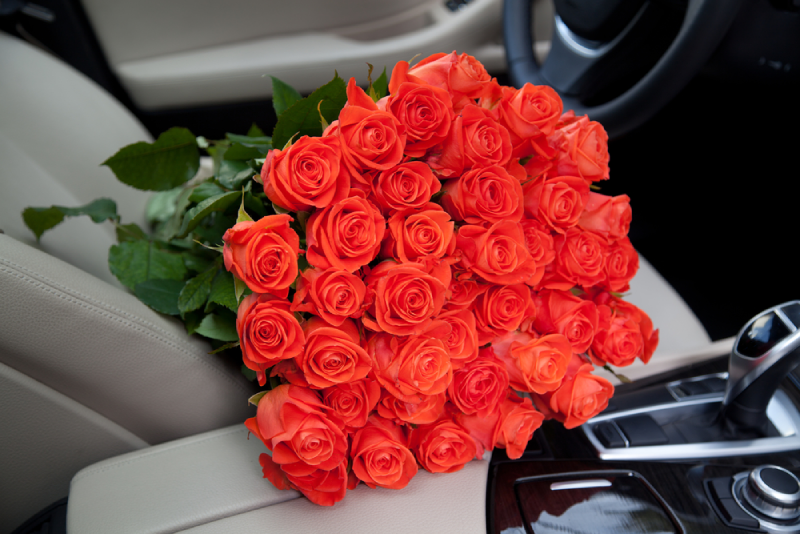 A large bouquet of vibrant orange roses lying across the light-colored car seat with the steering wheel in the background.