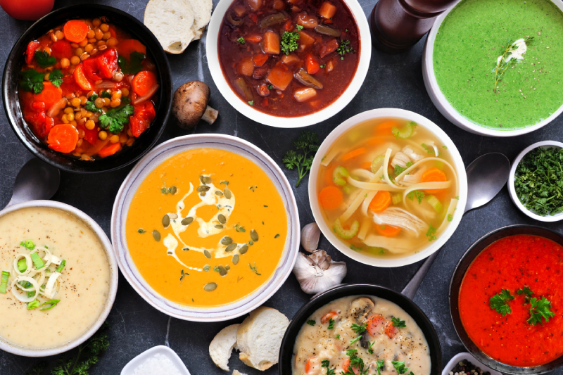 Overhead shot of 8 bowls of different colorful soups, including red, orange, green, and cream varieties.