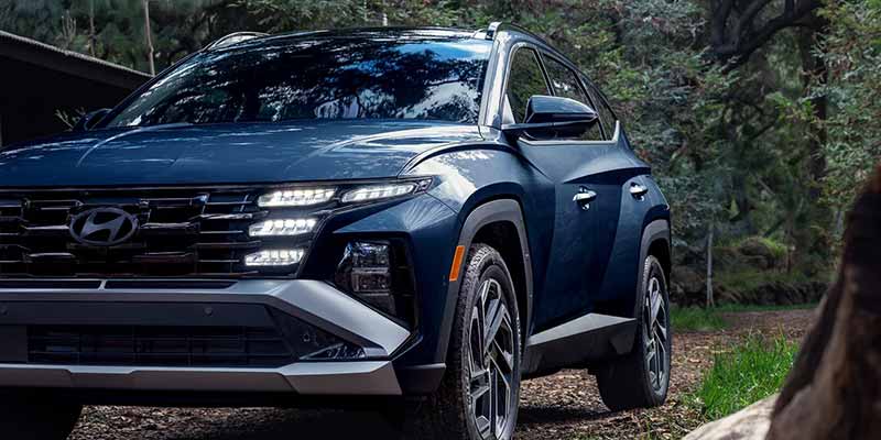 A sleek, dark blue Hyundai SUV with bright headlights, parked on a dirt path in front of green trees.