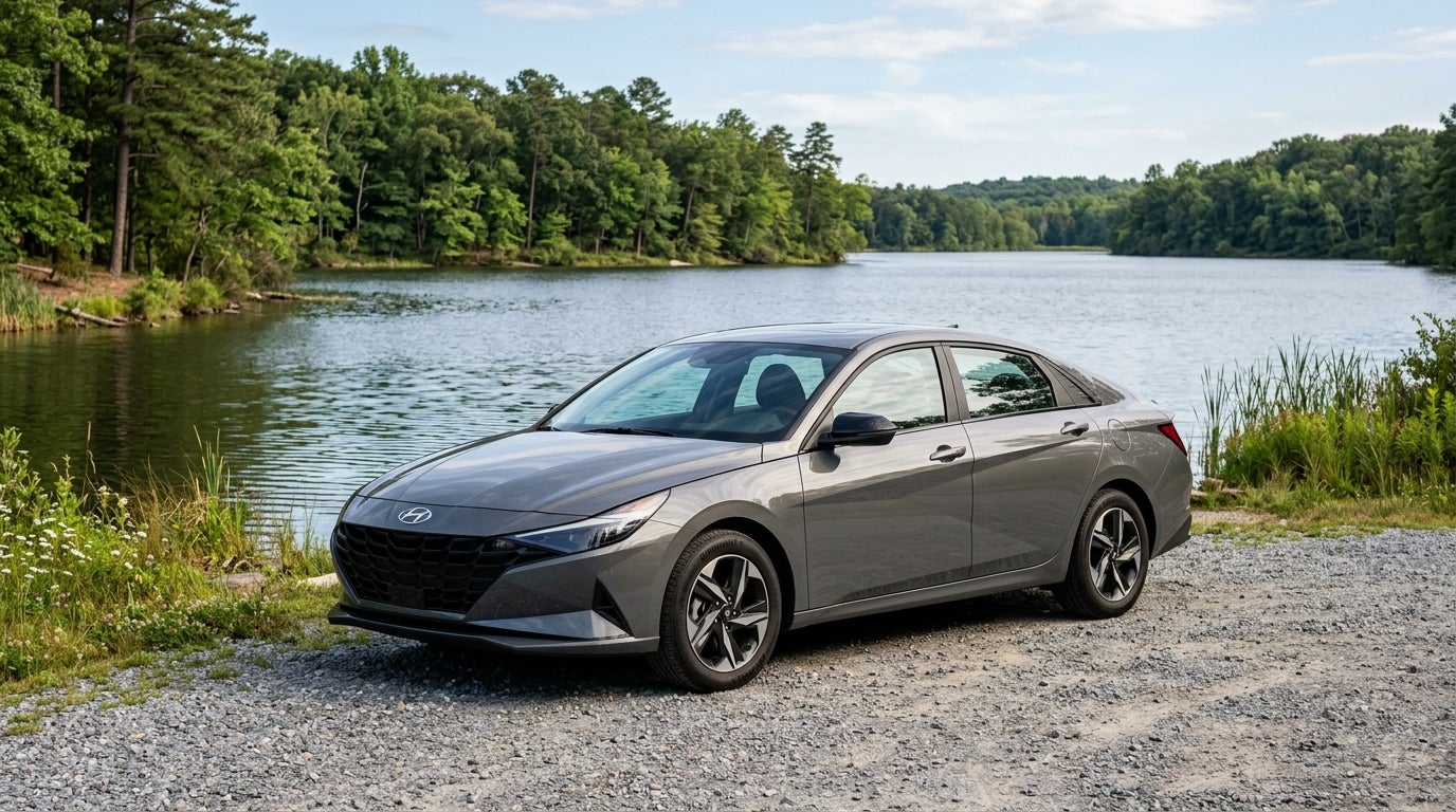 Grey Hyundai Elantra sedan parked on a gravel shore next to a calm lake surrounded by lush trees. (Representative image for illustration purposes only. Actual vehicle may vary based on trim and configuration.)
