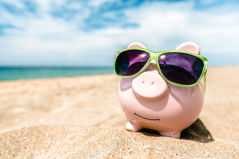 A pink piggy bank wearing green sunglasses on a sandy beach with the ocean and blue sky in the background.