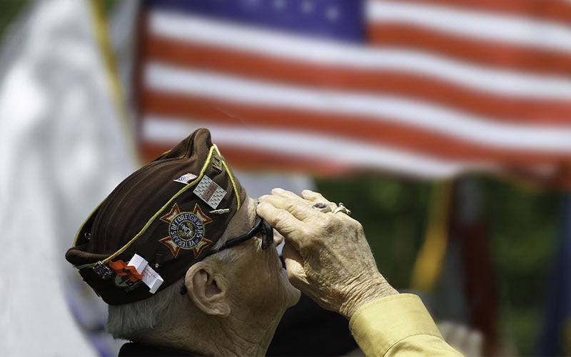 Elderly veteran in a brown cap saluting, with a blurred American flag in the background.