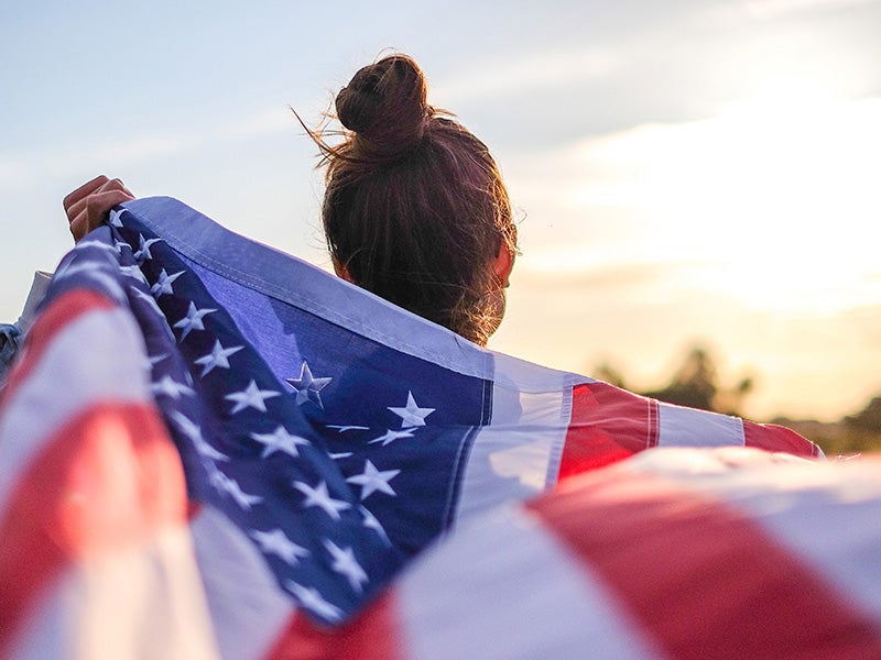 From behind, a person with a bun holds an American flag flowing over their shoulders against a bright sky.