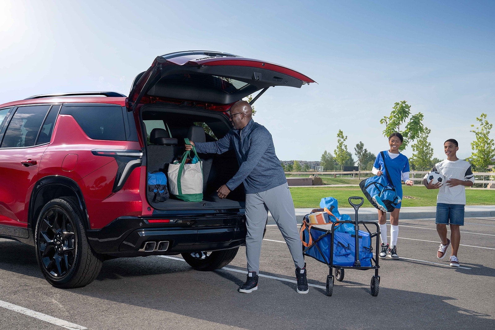 A man loads Chevy Traverse trunk while two kids with a soccer ball and sports bag.