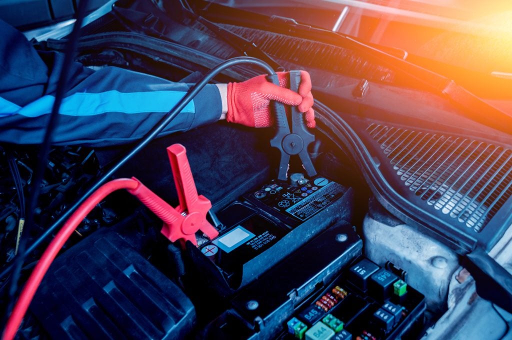 A mechanic wearing red gloves attaches jumper cables to a car battery under the hood.