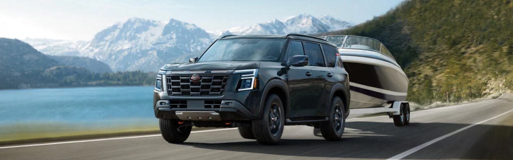 Black Nissan SUV towing a boat along a scenic lakeside road with mountains in the background.