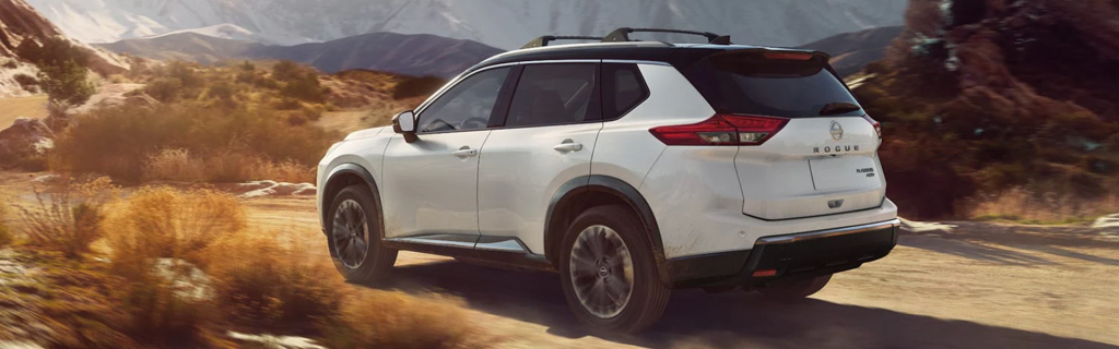 White Nissan Rogue driving on a dusty desert road with mountains in the background.