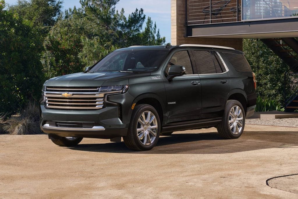 Brand new matte black 2023 Chevrolet Tahoe parked in a driveway facing left in front of a luxurious modern home.