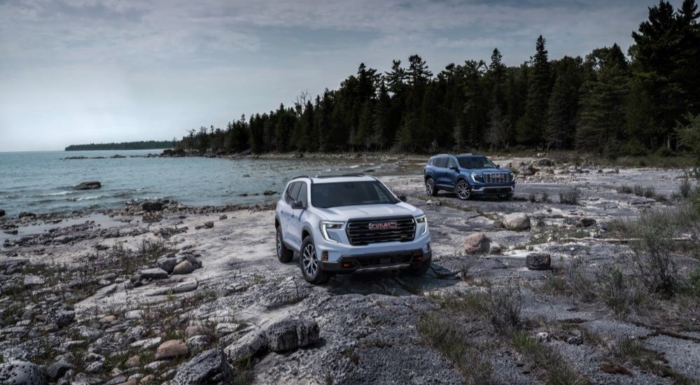 White 2024 GMC Acadia AT4 and a blue 2024 GMC Acadia Denali parked near a lake