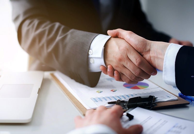 A closeup of two people in suits shaking hands over some finance forms.