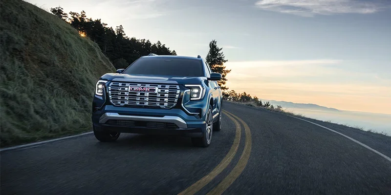 A blue 2026 GMC Terrain rounding a mountain curve with the blue sky sunrise in the background