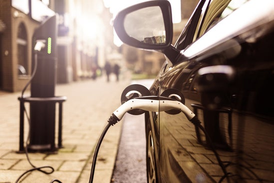A hand plugs a white and black charging connector into the side of a car. A blue charging cable extends from the connector.