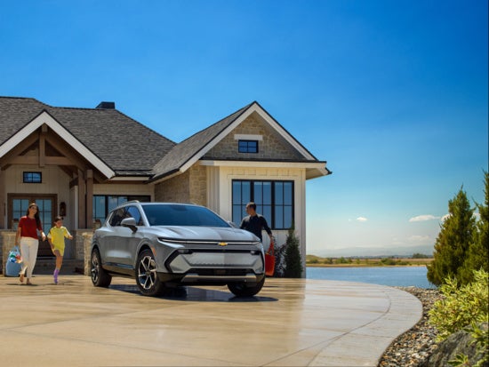 A silver Chevrolet Equinox EV SUV driving on a road, a lake and green trees in the background.