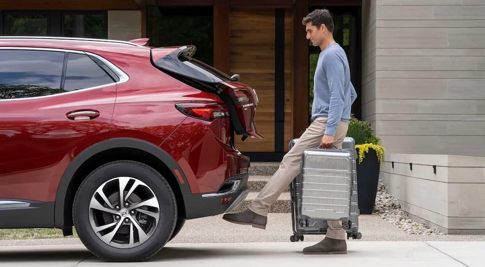 A man carrying luggage uses his foot to activate the power lift gate on a red 2023 Buick Envision.