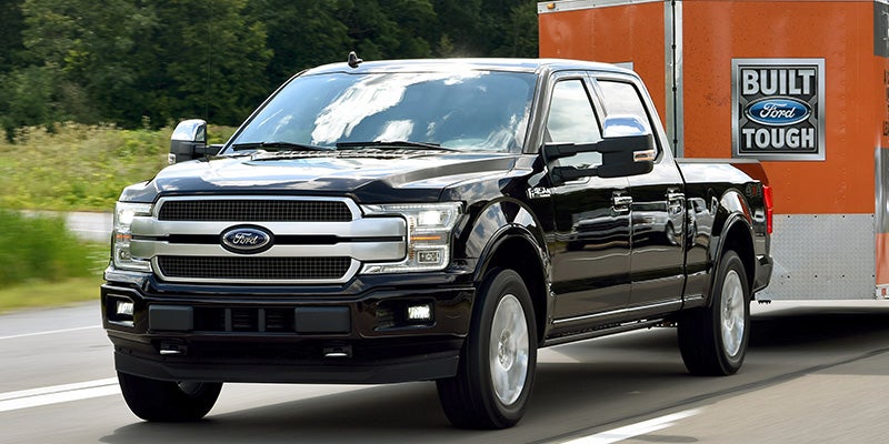 A black Ford F-150 SuperCrew truck towing an orange and silver cargo trailer on a highway.
