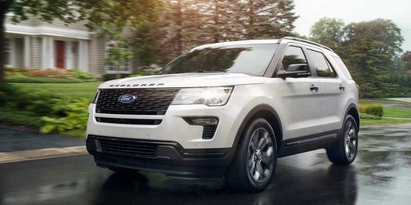 A white Ford Explorer SUV drives on a wet road, with a house and trees in the background.