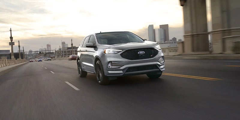 A silver Ford Edge ST drives on a multi-lane road towards the viewer, with a city skyline in the background.