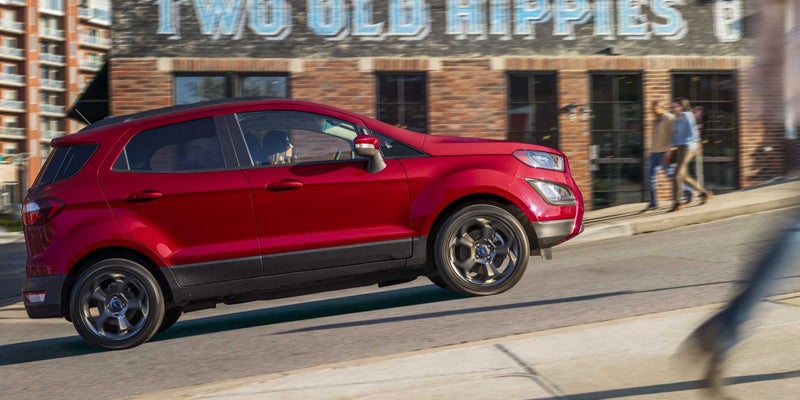 A red SUV drives up a street on a sunny day. A person is visible in the driver's seat. In the background left, there's a light brown brick building and a multi-story building. In the background right, two people walk on a sidewalk in front of a building with visible stores.