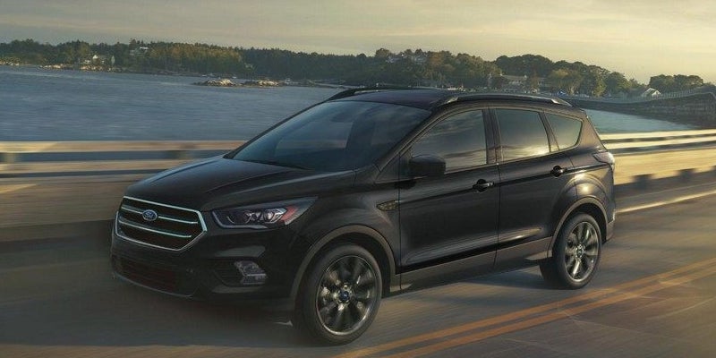 A black Ford SUV drives right on a highway, with a body of water and treelined coast in the background.
