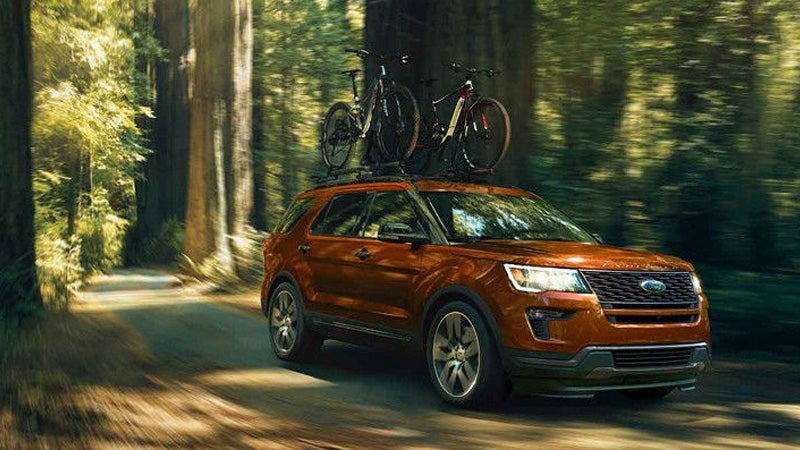 A copper SUV with two bicycles on its roof rack drives on a sun-dappled forest road.