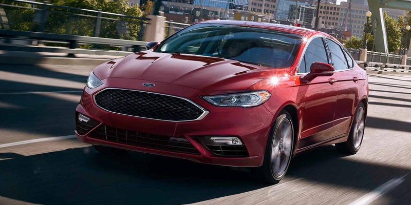 A shiny red Ford Fusion sedan drives toward the viewer on a bridge, with city buildings in the background.