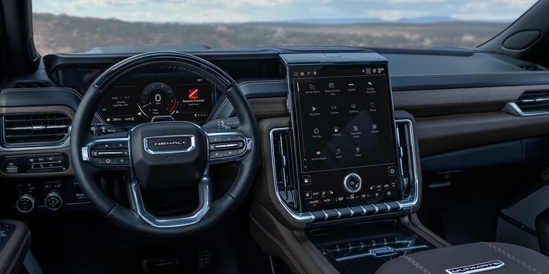 Interior view of a GMC Yukon Denali Ultimate dashboard featuring a leather-wrapped steering wheel, digital driver display, wood trim, and a large vertical infotainment touchscreen.
