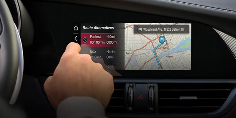 A hand interacts with a car's navigation screen showing route alternatives and a map of Detroit.