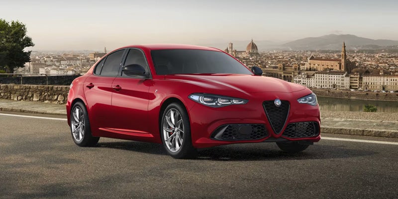 A red Alfa Romeo Giulia parked on a scenic road overlooking a city with a river and historic buildings.