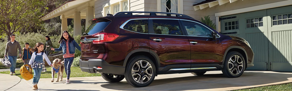 Side-rear view of a deep maroon Subaru Ascent parked in a residential driveway as a family with children and a dog walks toward it.