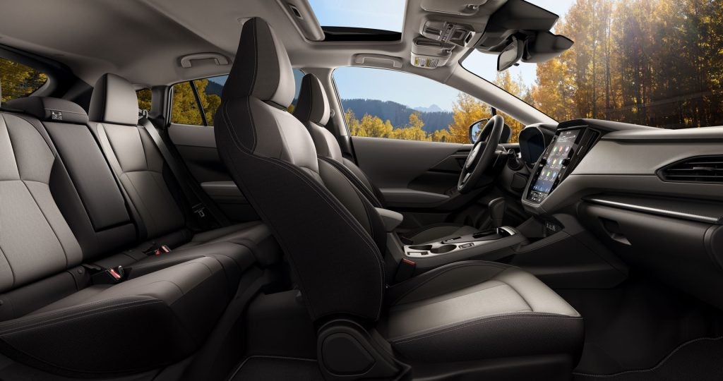 Interior view from the back seat of a vehicle featuring dark gray and tan cloth seats, a large vertical infotainment touchscreen, and an open sunroof looking out at autumn trees.