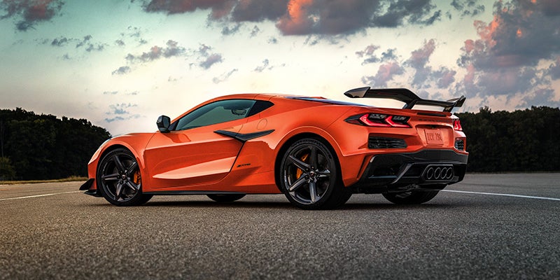 Rear view of an orange Chevrolet Corvette Z06 with a large rear wing parked on an asphalt road with trees and sky in the background.