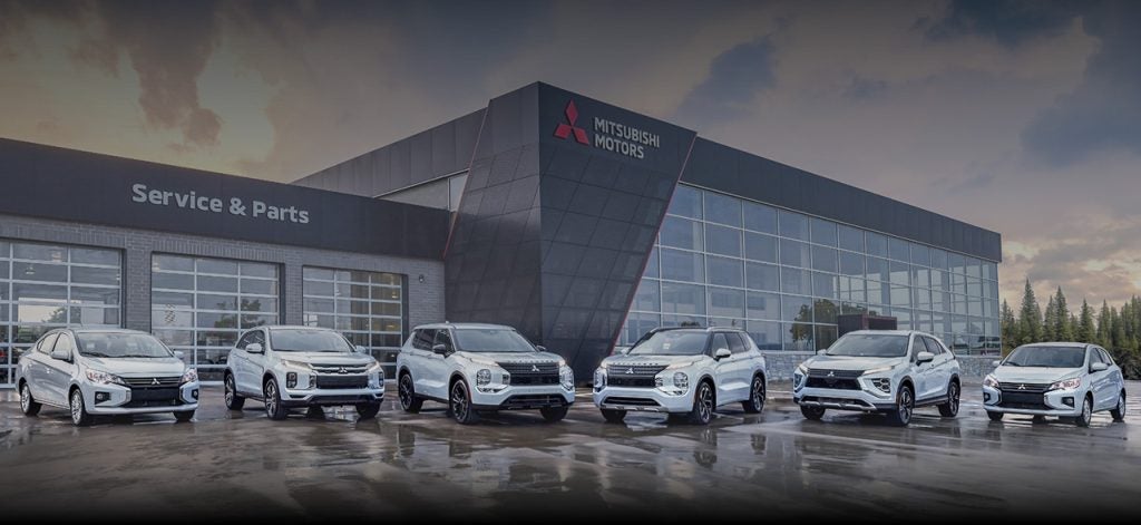 Six white Mitsubishi vehicles parked side-by-side on a wet asphalt surface in front of a modern Mitsubishi Motors dealership with a "Service &amp; Parts" wing under a cloudy evening sky.