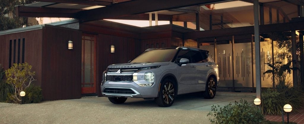 Front-three-quarter view of a light gray Mitsubishi Outlander parked under the modern carport of a wood-paneled mid-century house at dusk.