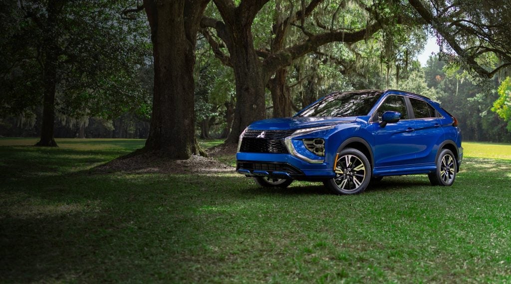 Front-three-quarter view of a bright blue 2026 Mitsubishi Eclipse Cross parked on a grassy field under large, shaded trees.