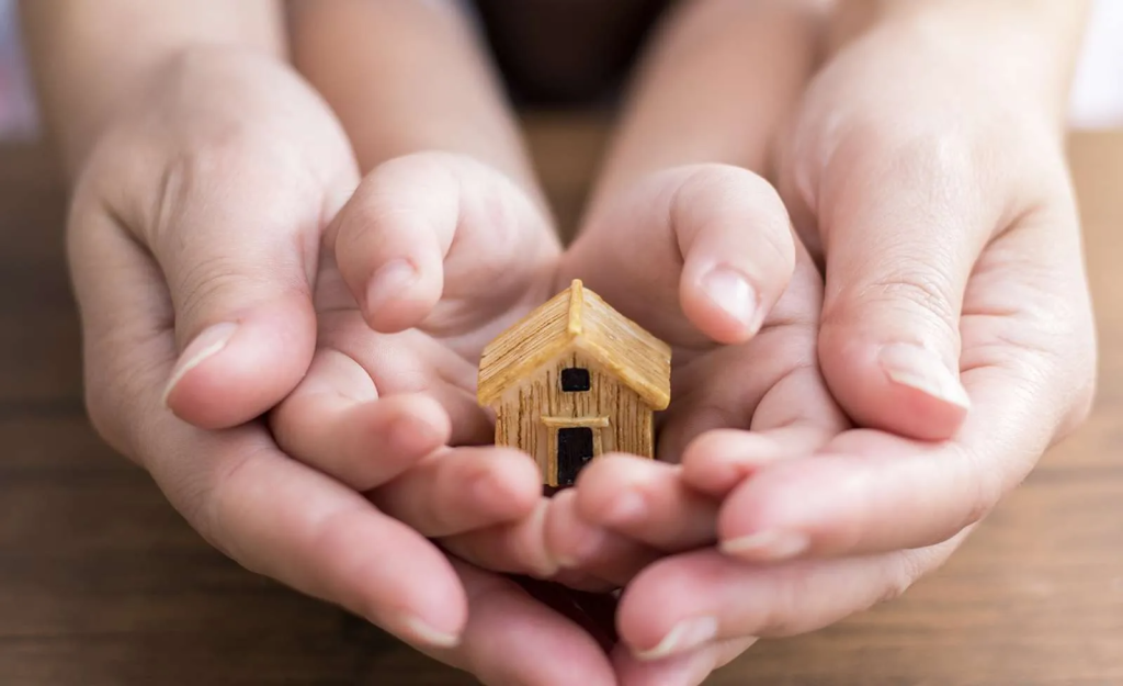 a pair of adult hands holding a pair of child hands holding a wooden model of a house representing Adding Your Child to the Title of Your Home