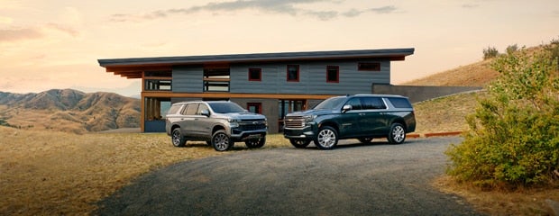 Two 2023 Chevrolet Suburban SUVs parked in front of a residential house on a sunny day.
