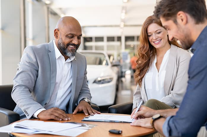 Couple signing paperwork while car dealer holds out a set of keys.