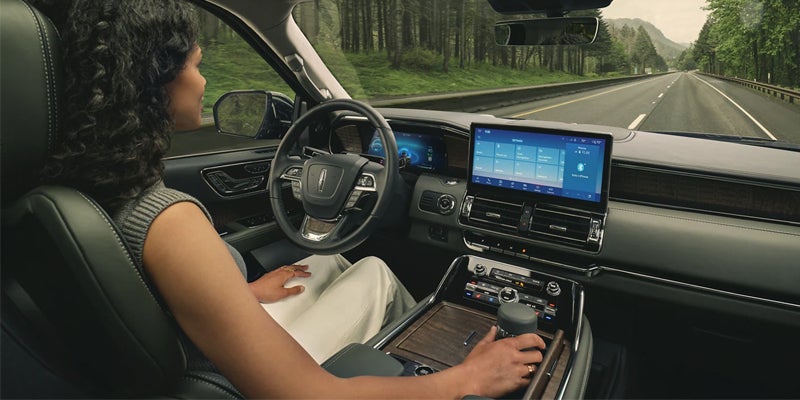 A woman driving a Lincoln vehicle on a highway through a forest.