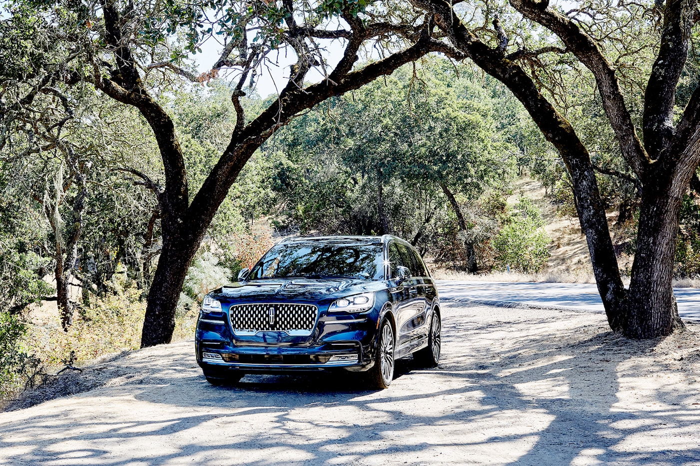 Blue Lincoln Aviator driving along a sunlit, winding forest road.