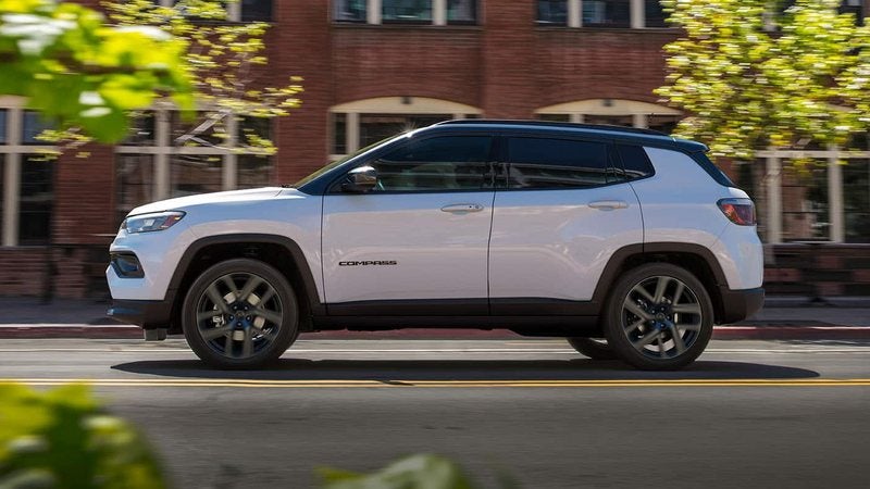 White 2026 Jeep Compass driving past brick storefronts; Jeep Compass specials at Ross Downing CDJR of Mobile, AL