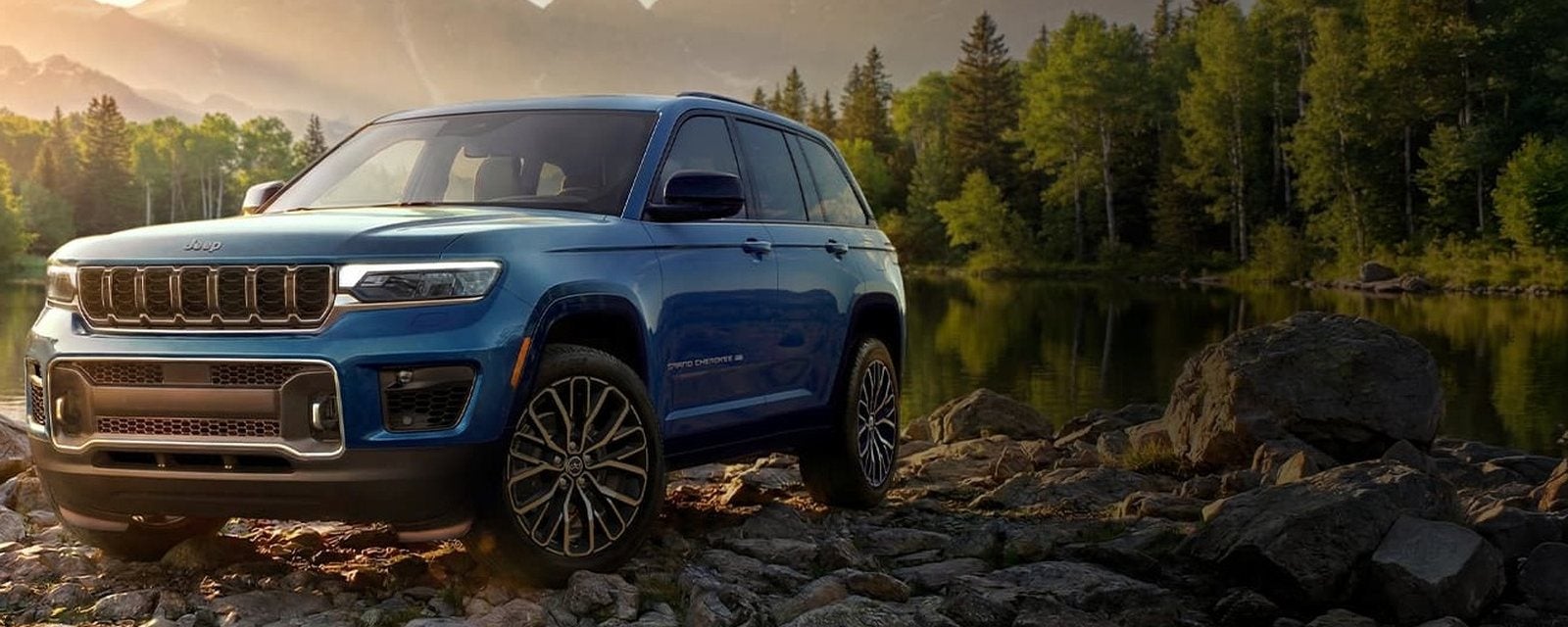 Blue 2027 Jeep Grand Cherokee Overland on a rocky lakeshore in golden light; new Grand Cherokees for sale at Ross Downing CDJR of Mobile, AL