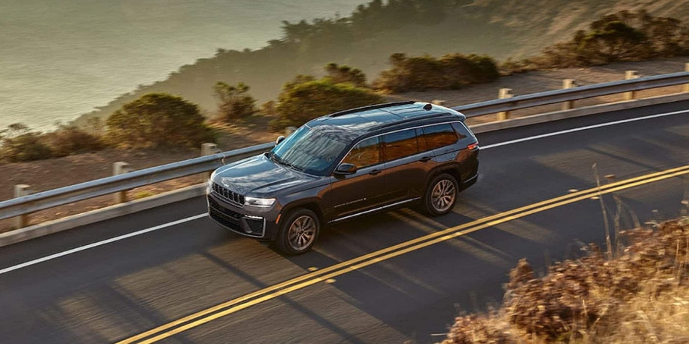 Gray 2026 Jeep Grand Cherokee L Summit driving a coastal highway at sunset, for sale at Ross Downing CDJR of Mobile, AL