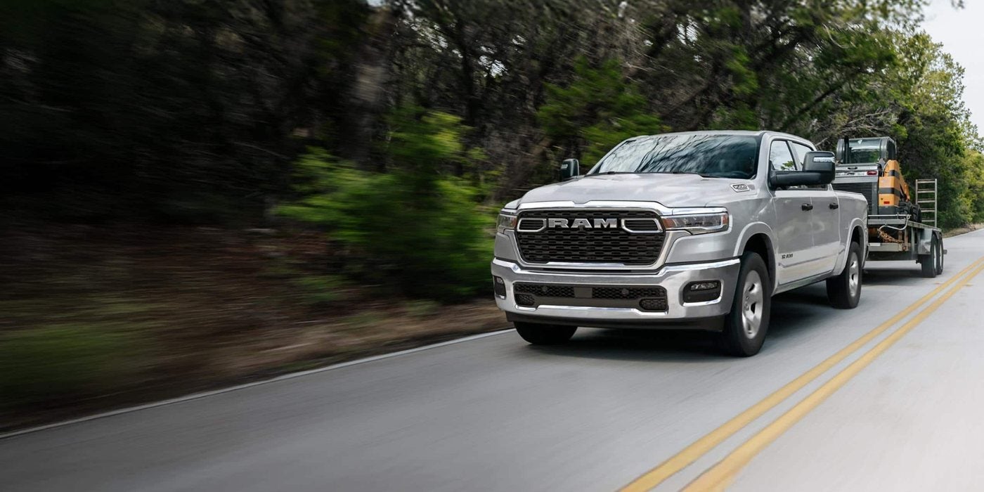 Silver 2026 Ram 1500 towing an equipment trailer on a two-lane road, for sale at Ross Downing CDJR of Mobile, AL
